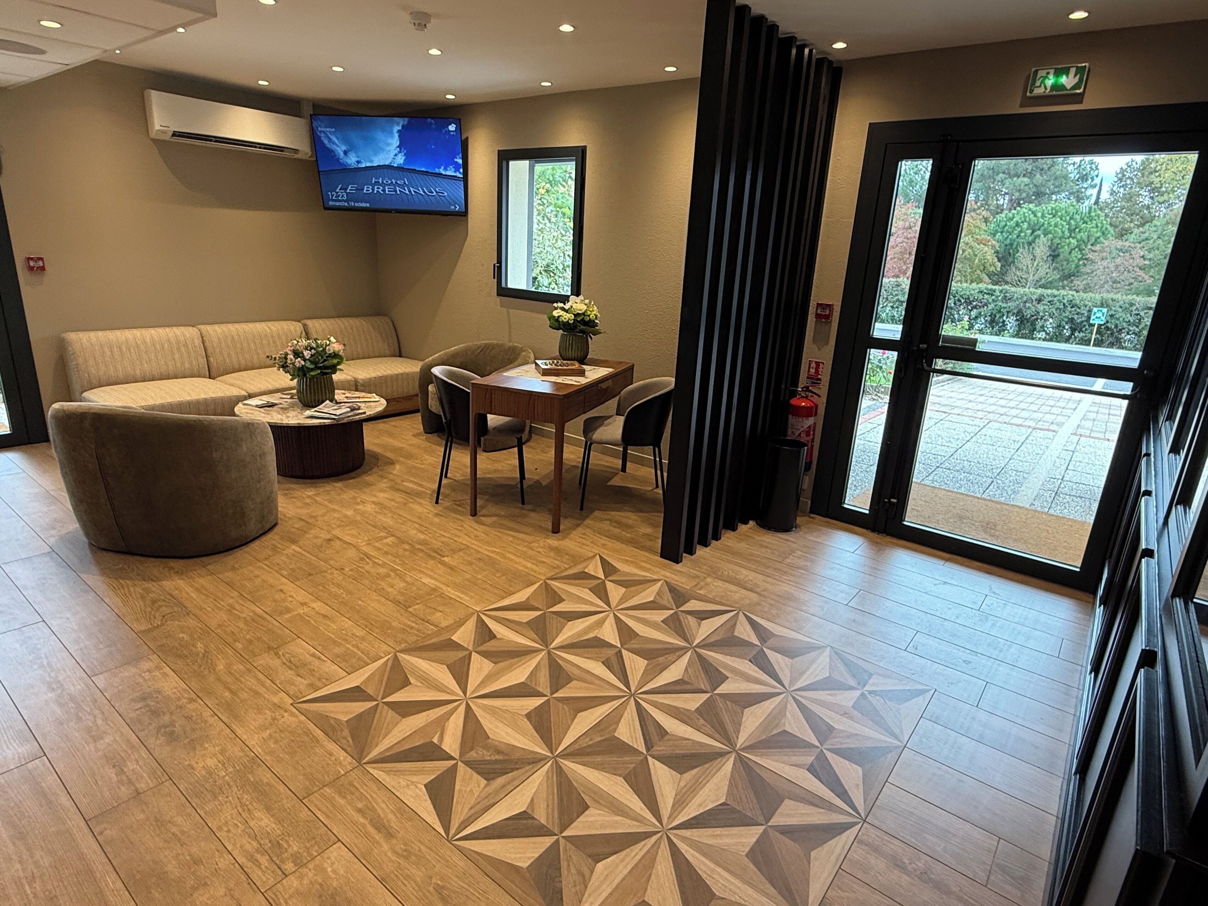Un hall de hotel con un sofá, una mesa con un florero, un suelo estampado, una pantalla de televisión y una puerta que da al exterior.