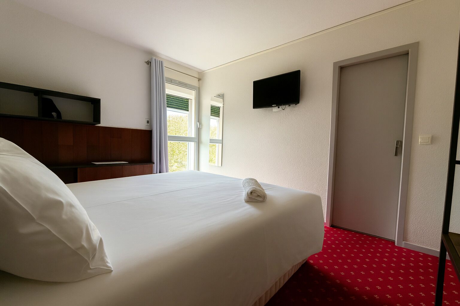 Een hotelkamer met een bed, een aan de muur bevestigde televisie, een deur en een raam met gordijnen.