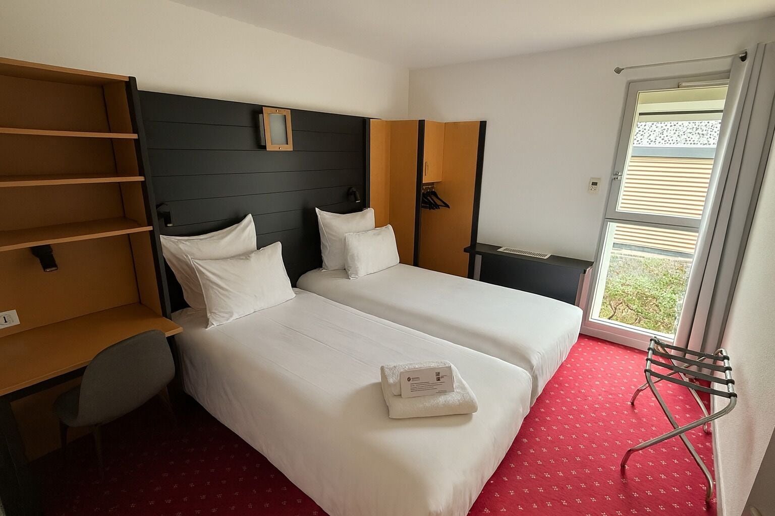 Quarto de hotel com duas camas, um guarda-roupa, uma escrivaninha e uma janela com persianas.