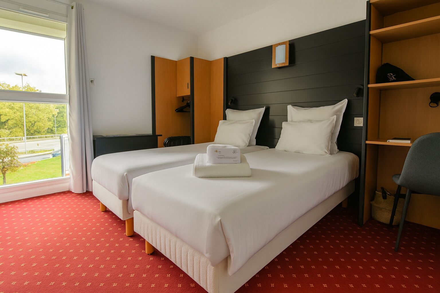Quarto de hotel com duas camas, tapete vermelho, cabeceira de madeira, uma mesa de trabalho e vista para uma paisagem verde.