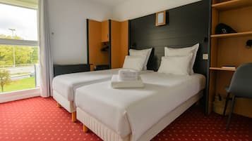 A hotel room with two beds, a red carpet, a wooden headboard, a desk, and a view of a green landscape.