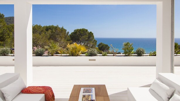 Un espace extérieur moderne, aménagé avec des canapés blancs et une table en bois, offrant une vue panoramique sur la mer et une végétation luxuriante.