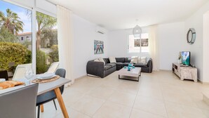 A spacious living room with a large sofa, a dining table with chairs, and a television on a wooden stand. - Green Bay Villa #14 (Paralimni)
