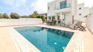 A modern pool area with a rectangular pool, lounge chairs, and a dining table. - Green Bay Villa #14 (Paralimni)