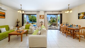 A spacious living area with a green sofa, a beige sofa, and a dining table set for four. Large windows offer a view of the pool and palm trees. - Villa Greco Mare #5 (Paralimni)