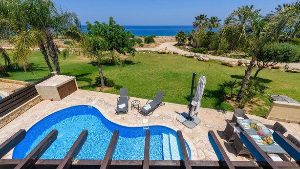 A swimming pool with lounge chairs and a table, surrounded by a landscaped garden and a view of the ocean. - Villa Greco Mare #5 (Paralimni)