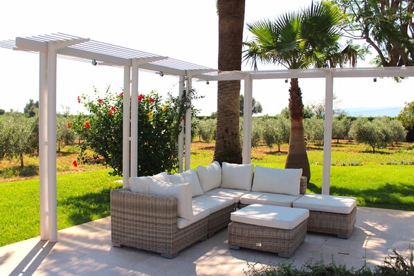 A covered outdoor seating area with a white pergola, wicker furniture, and a view of an olive grove.