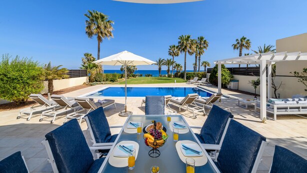 A poolside dining area with a view of the sea, surrounded by palm trees and lounge chairs. - Royal Beachfront Villa (Paralimni)