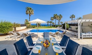 A poolside dining area with a view of the sea, surrounded by palm trees and lounge chairs. - Royal Beachfront Villa (Paralimni)