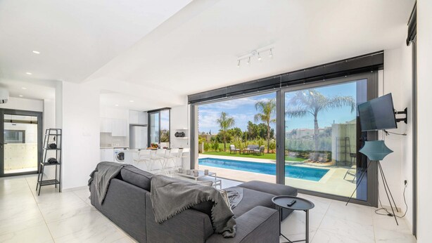 Ein modernes Wohnzimmer mit einem grauen Sofa, einem kleinen Tisch und einem Fernseher. Ein großes Fenster bietet Blick auf einen Swimmingpool und Palmen.