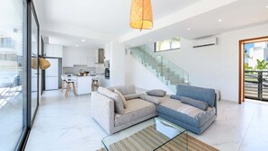 A modern living room with a sofa, a glass coffee table, and a hanging light fixture. - By The Beach - White Coral Villa (Paralimni)