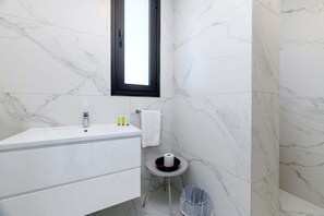 Un baño moderno con un lavabo de mármol blanco, una mesita redonda con artículos de aseo personal y un cubo de basura.