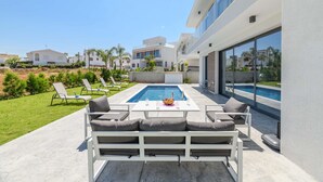 A modern outdoor pool area with a white pool table, lounge chairs, and a sofa. - By The Beach - White Coral Villa (Paralimni)