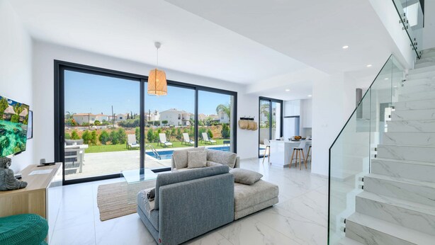A modern living room with a large sliding glass door leading to a pool area, a grey sofa, and a staircase with marble steps. - By The Beach - White Coral Villa (Paralimni)