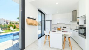 A modern kitchen with white cabinets, a central island, and a view of a swimming pool. - By The Beach - White Coral Villa (Paralimni)