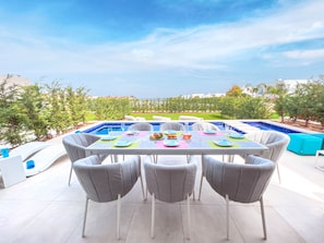 A dining area with a pool view, featuring a table set for four with chairs, a blue pool, and a view of a landscaped garden. - Azure Seaview - Jade Villa (Paralimni)