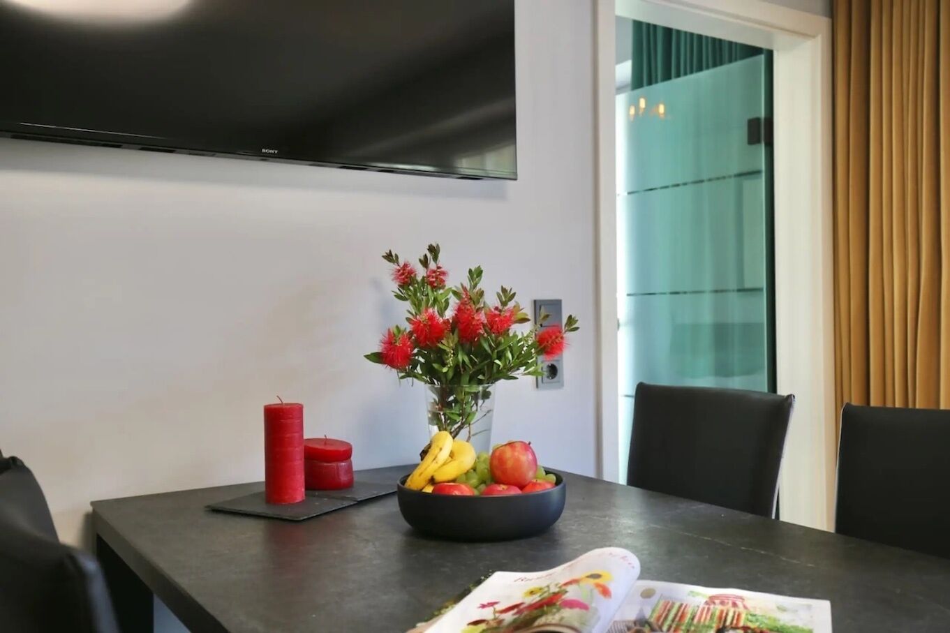 Ett modernt matområde med en platt-TV som hänger på väggen, ett bord med en vas med blommor, en skål med frukt och en tidning.