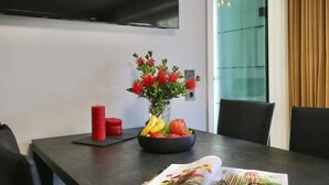 A modern dining area with a flat-screen TV mounted on the wall, a table with a vase of flowers, a bowl of fruit, and a magazine. - Island jewel in the Lindenhof with south-facing terrace (Wangerooge)