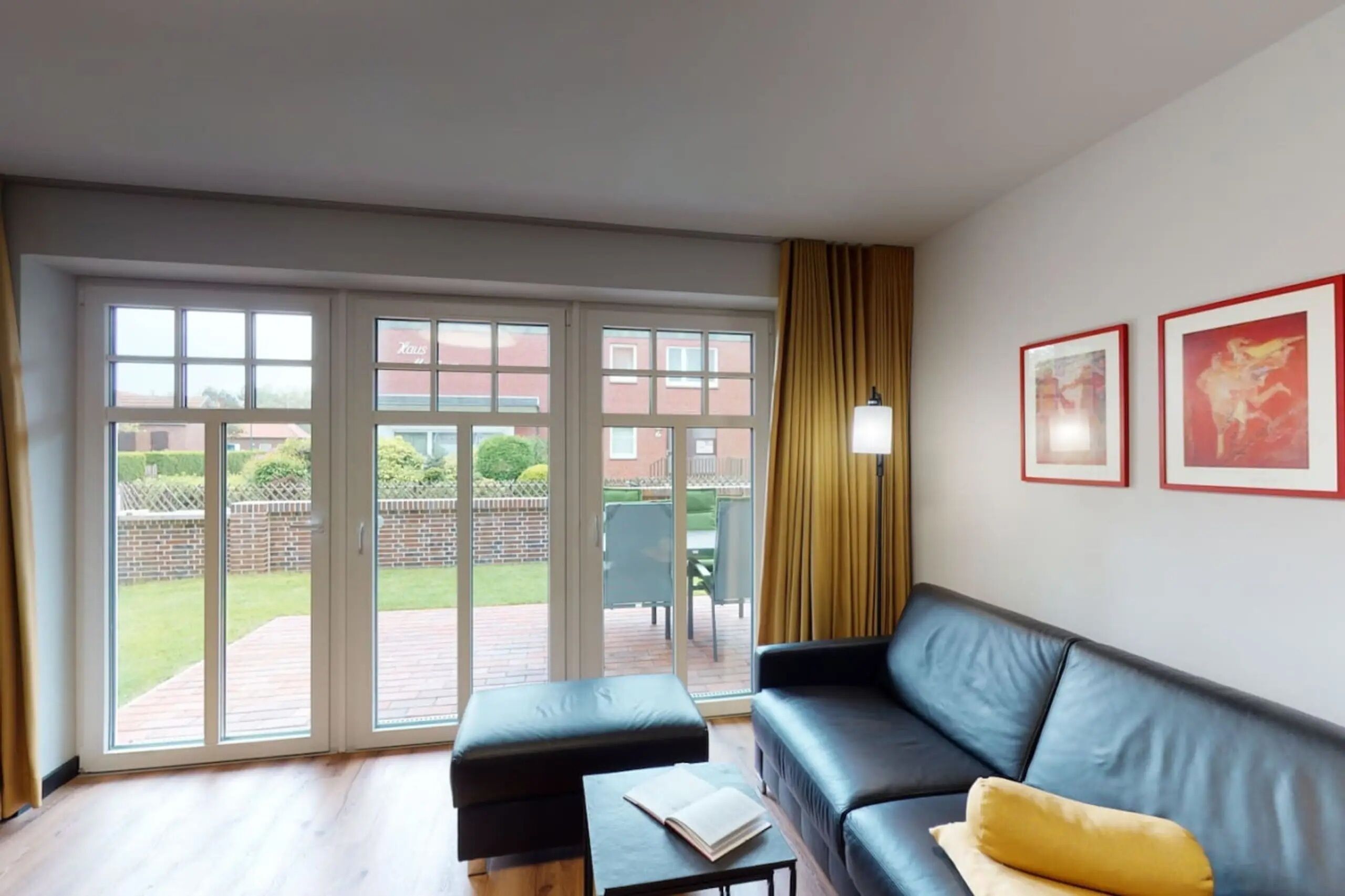 Un salon avec un canapé en cuir noir, une petite table sur laquelle repose un livre, et une porte-fenêtre coulissante donnant sur une terrasse extérieure avec une chaise et une table.