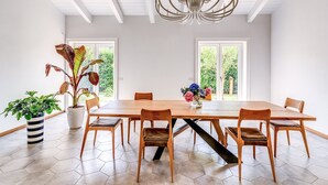 Un comedor con una gran mesa de madera, seis sillas, una araña, plantas en macetas y ventanas amplias.