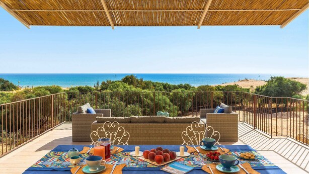 Tavola apparecchiata per la colazione con vista sul mare, mobili in vimini e tovaglia azzurra.