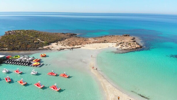 Eine Luftaufnahme eines Strandes mit klarem türkisblauem Wasser, einem Felsen und mehreren kleinen Booten sowie Luftmatratzen, die an der Küste liegen.