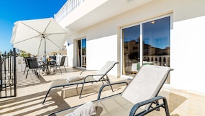 Ein sonniger Sitzbereich im Freien mit Liegestühlen und einem Tisch, einem weißen Sonnenschirm und einer Glastür zu einer überdachten Terrasse.