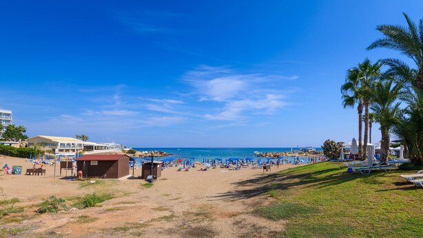 A beach with palm trees, beach umbrellas, and a building with a red roof. - Pernera Blue Villa (Paralimni)