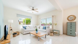 A spacious living room with a white sofa, a wooden coffee table, a ceiling fan, and a view of the outdoors through large windows. - Pernera Blue Villa (Paralimni)