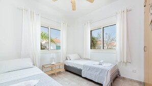 A hotel room with two beds, a small table, and a window with curtains. - Pernera Blue Villa (Paralimni)