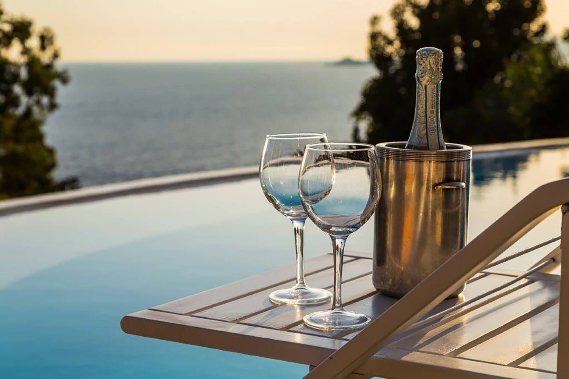 Ett bord med två vinglas och en champagneflaska, placerat vid poolkanten med utsikt över havet.