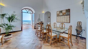 Ein Essbereich mit einem Glastisch und Holzstühlen, Blick auf das Meer durch einen offenen Bogen, und eine Wanddekoration.