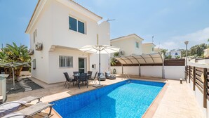 A swimming pool with lounge chairs and a table, surrounded by a white building with a tiled roof and a covered area. - Villa Amesta (Paralimni)