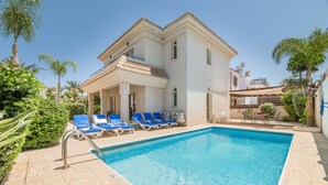 A two-story building with a swimming pool, blue lounge chairs, and palm trees. - Villa Lindo (Paralimni)