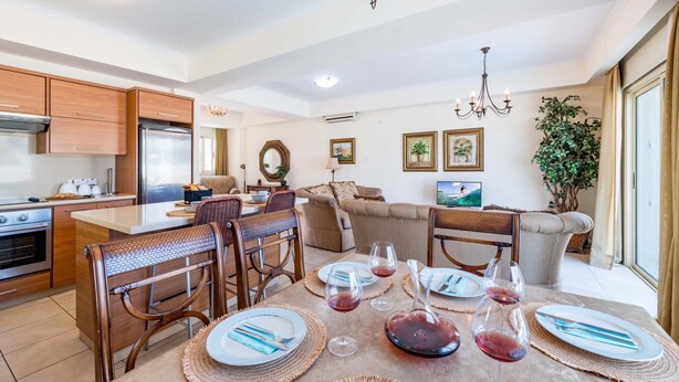 A modern kitchen with wooden cabinets, a dining area with a table set for four, and a living area with a sofa and a large plant. - Villa Jacqueline (Paralimni)