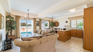 A spacious living area with a sofa, a dining table, and a kitchen area with wooden cabinets and a sink. - Villa Jacqueline (Paralimni)