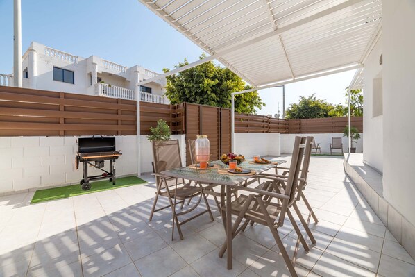 Een terras buiten met een tafel, stoelen, een barbecue en een overdekt gedeelte.