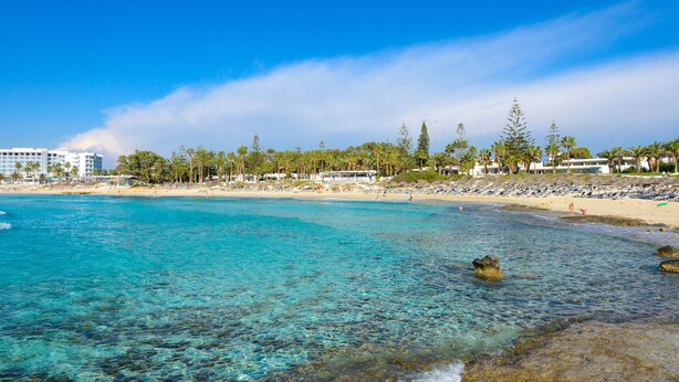 A beach with clear turquoise water, a sandy shore, and a row of beachside buildings with umbrellas. - Napa Centro Apartment (Ayia Napa)