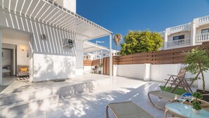 A modern outdoor patio with a covered seating area, a small table with a plant, and a view of a white building with balconies. - Napa Centro Apartment (Ayia Napa)