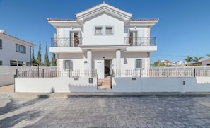A two-story house with a white facade, a balcony on the upper floor, and a gated entrance. - Nemea Serenity Blue (Ayia Napa)