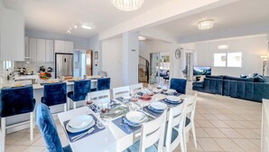 A modern kitchen with white cabinets, a dining area with a table set for four, and a living area with a dark blue sofa and a TV. - Nemea Serenity Blue (Ayia Napa)