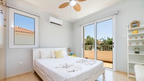 A bedroom with a bed, a ceiling fan, a window, an air conditioning unit, a balcony with a view, and a shelf with jars. - Villa Kohili (Paralimni)