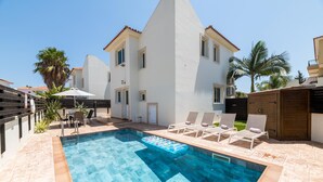 A swimming pool with lounge chairs and a wooden deck, surrounded by a white building with a tiled roof. - Villa Kohili (Paralimni)