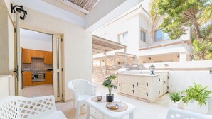 A bright outdoor patio with white furniture, a small table with a potted plant, and a view of the kitchen area. - Anthorina Greco Villa #10 (Paralimni)