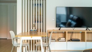 Egy modern nappali, melyben egy fa étkezőasztal, fehér székek, egy fa talapzatra helyezett televízió és egy könyvespolc található.