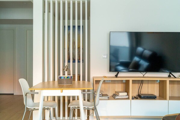 Una sala de estar moderna con una mesa de comedor de madera, sillas blancas, un televisor sobre un mueble de madera y una estantería.