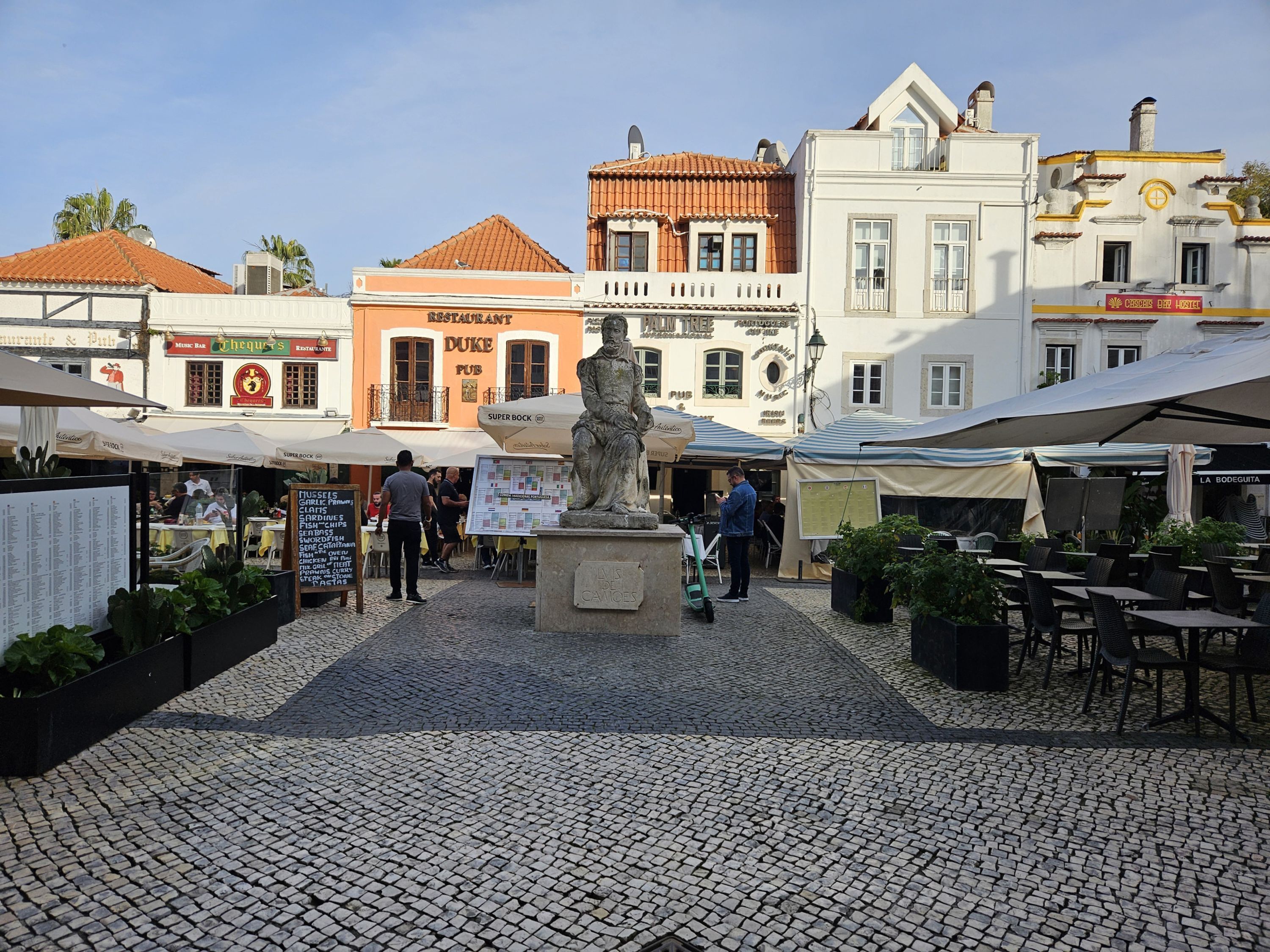 Une place pavée avec des tables en plein air, une statue et divers bâtiments portant des enseignes de restaurants et d’un pub.