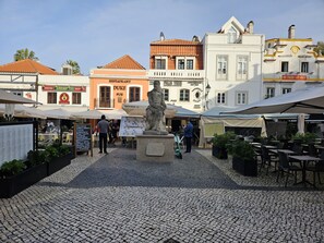 Une place pavée avec des tables en plein air, une statue et divers bâtiments portant des enseignes de restaurants et d’un pub.