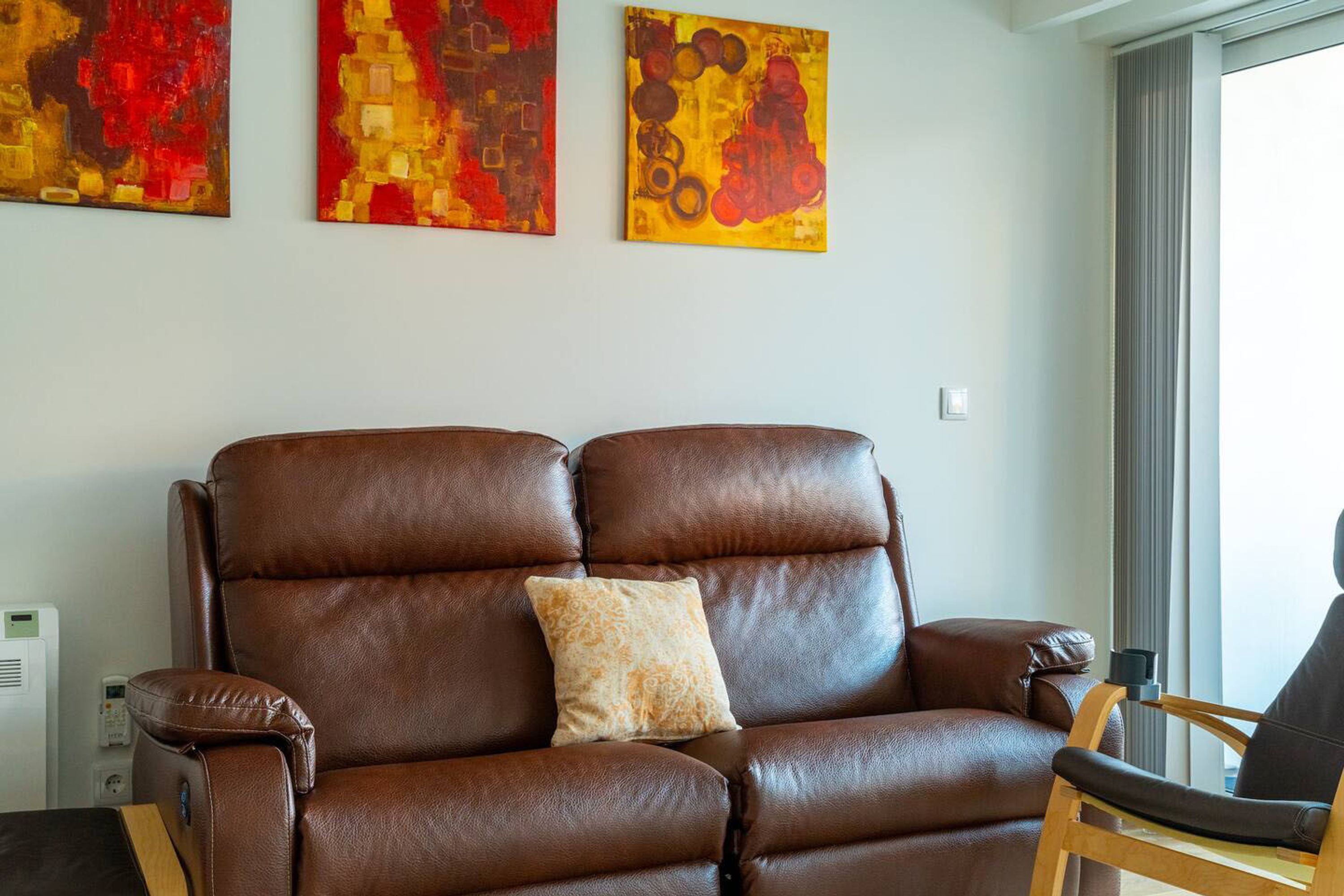 Un canapé en cuir marron agrémenté d’un coussin à motifs, une chaise en bois et trois tableaux abstraits accrochés au mur.