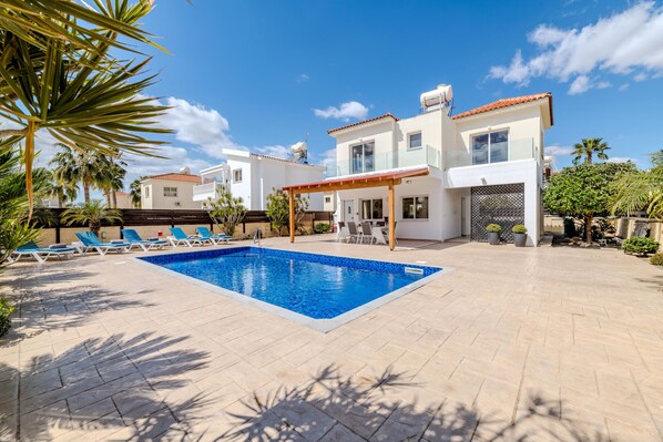Una piscina con sillas de descanso, un porche cubierto y una casa de dos pisos con tejado de baldosas.